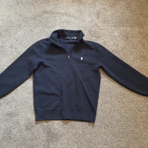 Mörkblå half zip tröja Polo Ralph Lauren - Snygg mörkblå tröja från Polo Ralph Lauren med klassisk half zip och broderad logga på bröstet. Tröjan har långärmade ärmar och stårkrage, perfekt för dig som gillar stilren och enkel design. Materialet är mjukt och skönt, troligtvis bomull. Priset kan sänkas vid snabba affärer