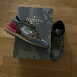 Valentino Rockrunner camo sneakers - Säljer ett par Valentino Rockrunner sneakers med camouflagemönster i grönt, beige och grått. Skorna har detaljer i mocka och skinn, samt en cool röd häl med nitar. Klassisk snörning och rund tå. Perfekt för dig som vill sticka ut med lyxiga street vibes.
