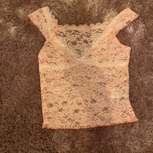 Ljusrosa spetsig linnetopp - Supersöt ljusrosa linnetopp i spets med v-ringning och små volangdetaljer längs axelbanden. Toppen är ärmlös och har en liten rosett framtill. Perfekt för dig som gillar romantisk och feminin stil.