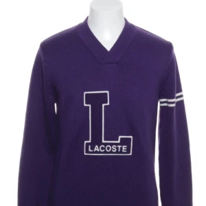 Lila stickad Lacoste tröja med V-ringning - Snygg lila stickad tröja från Lacoste med V-ringad hals och stor L-logga framtill. Tröjan har långa ärmar med vita ränder på ena armen och ribbade muddar. Perfekt för dig som gillar sportig och retro stil.