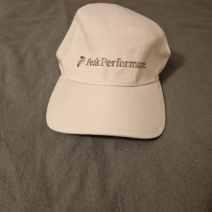 Oanvänd Peak Performance keps S/M - Oanvänd. Snygg vit keps från Peak Performance, modell Kinloch Cap. Klassisk form med böjd skärm och broderad logga i guld på framsidan. Insidan har svart svettband med logotyp. Tillverkad i bomull, perfekt för dig som gillar stilren sportig look.