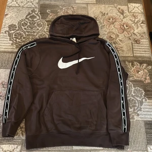 Brun Nike hoodie med vita detaljer byxor ingår i priset - Säljer en brun hoodie från Nike med stor vit swoosh-logga på bröstet och vita loggor längs ärmarna. Tröjan har huva med snörning, känguruficka och ribbade muddar. Perfekt för chill eller sport. Materialet är mjuk bomull/polyesterblandning och passformen är normal.
