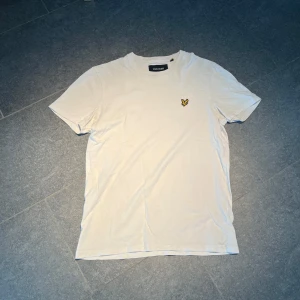 Vit t-shirt från Lyle & Scott - Vit t-shirt från Lyle & Scott med klassisk rund hals och korta ärmar. På bröstet sitter den ikoniska broderade gula fågeln. T-shirten är gjord i mjuk bomull och har en clean, stilren look som funkar till jeans eller shorts.