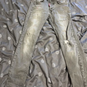 Grå Dsquared2 jeans med slitningar - Säljer ett par grå jeans från Dsquared2 med coola slitningar och färgstänk på framsidan. Modellen har en rak passform och klassisk femficksdesign. Jeansens material är bomull och de har en snygg Dsquared2-patch bak i midjan. Perfekt för dig som gillar streetstyle.