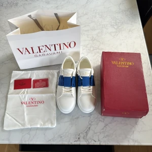 Valentino Garavani Open sneakers vit/blå - Säljer ett par Valentino Garavani Open sneakers i vitt skinn med en snygg blå läderdetalj över snörningen. Skorna har rund tå, platt sula och ikoniska nitar på hälen. Kommer med originalkartong, dustbag och äkthetskort.