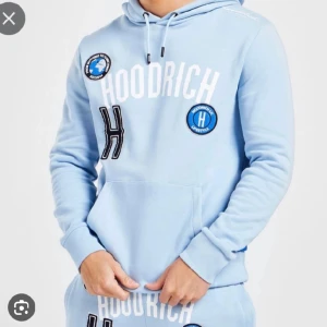 Ljusblå hoodie från Hoodrich - Säljer en ljusblå hoodie från Hoodrich med vita och svarta tryck samt broderade patches på bröstet. Tröjan har huva med snörning och en stor magficka. Perfekt för dig som gillar streetwear och vill sticka ut med coola detaljer.