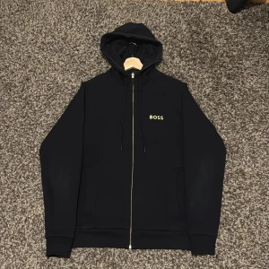 Svart hoodie med guldfärgad dragkedja - BOSS - Svart hoodie från BOSS med guldfärgad dragkedja. Har använts men inga direkt tecken på att den har använts eller är skadad osv. Säljer denna då den har blivit stor för mig. Önskas fler bilder så är det bara att hojta till. Kvitto finns! Ny pris cirka 1200kr.