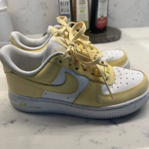 Nike Air Force 1 sneakers i ljusgul, exklusiva - Nike Air Force 1 sneakers i en fräsch ljusgul och vit färgkombination. Klassisk låg modell med snörning, perforerad tå och platt sula. Swoosh-logga på sidan och AIR-detalj på sulan. Tillverkade i skinn med textilfoder för extra komfort. Perfekt för dig som gillar stilrena och färgglada kicks.