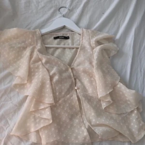 Prickig blus med volanger från Bik Bok - Säljer en ljusbeige blus från Bik Bok med prickigt mönster och pärlknappar framtill. Blusen har volangdetaljer och korta, luftiga ärmar. Stl xs💕