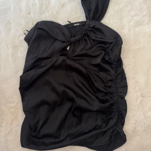 Svart oneshoulder topp från Bik Bok - Snygg svart oneshoulder topp från Bik Bok i glansigt, mjukt material. Toppen har en cool knytning vid axeln och rynkade detaljer längs sidan som ger en unik look. Perfekt för dig som vill sticka ut med en trendig och stilren outfit.