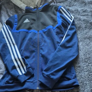 Adidas blå vindjacka med dragkedja - Snygg blå och svart vindjacka från Adidas med klassiska vita ränder längs ärmarna och logga på bröstet. Jackan har hel dragkedja framtill, ribbade muddar och sportig look. Perfekt för dig som gillar streetstyle och sportmode. Original priset är 1300kr.