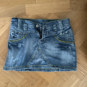 Blå jeanskjol från CB Jeans låg midja  - Snygg blå jeanskjol från CB Jeans med coola detaljer som kontrasterande gula sömmar, fickor både fram och bak samt bälteshällor. Kjolen har en klassisk kort passform och dragkedja framtill. Perfekt för dig som gillar streetstyle och denim.
