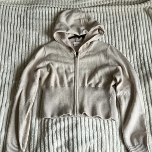 Beige croppad hoodie från Gina Tricot - Säljer en beige croppad hoodie från Gina Tricot med dragkedja framtill och huva. Tröjan har långa ärmar och bred ribbad kant vid midjan och ärmsluten. 