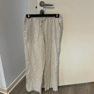Beige randiga linnebyxor med snörning - Snygga beige linnebyxor med vita vertikala ränder och avslappnad passform. Byxorna har elastisk midja med snörning och raka ben, perfekta för chill dagar. Luftigt material som känns skönt mot huden och ger en fräsch vibe.