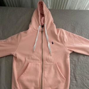 Ljusrosa hoodie från Polo Ralph Lauren - Säljer en ljusrosa hoodie från Polo Ralph Lauren i storlek M. Tröjan har huva med vita snören, dragkedja framtill och en broderad mörkblå logga på bröstet. Mjuk insida i fleece och praktisk känguruficka. Perfekt för chill dagar och snygg streetstyle.
