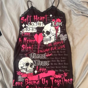 Svart kort klänning med döskallar och text - Cool svart kort klänning med tunna axelband och spetsdetaljer vid halsringning och nederkant. Framsidan har ett stort tryck med vita döskallar, rosa hjärtan och edgy text i rosa och vitt. Klänning men går nog att använda som nattlinne också. Aldrig använd i storlek XL/42 men passar även L. Säljer för 50kr
