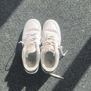 Nike Air Force 1 vita sneakers storlek 41 - Säljer ett par klassiska Nike Air Force 1 sneakers i vitt med svart swoosh. Skorna har snörning, rund tå och platt sula. Ovandelen är i präglat skinn och de har den ikoniska Air-sulan för extra komfort. Perfekt för dig som gillar streetstyle och vill ha en tidlös look.