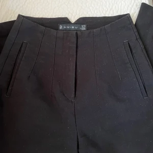 Svarta kostymbyxor från Amisu - Snygga svarta kostymbyxor från Amisu med hög midja och raka ben. Byxorna har stilrena fickor framtill och en cool V-detalj i midjan. Perfekta för dig som gillar en clean och klassisk look. Materialet känns mjukt och har en lätt glansig finish.