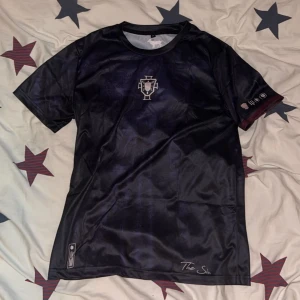 Svart fotbollströja  - Snygg svart fotbollströja från The Siu med diskret mönster och tryckt nummer 7 på ryggen. Tröjan har korta ärmar, rund hals och är gjord i ett glansigt, lätt polyestermaterial. Unik logga på bröstet och get-symbol vid nacken. Perfekt för dig som gillar stilren design.