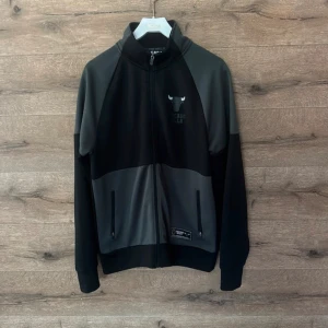 Chicago Bulls ziptröja NBA svart/grå - Snygg ziptröja från NBA med Chicago Bulls-logga både fram och bak. Svart och grå färgblock, hög krage och hel dragkedja. Två fickor med dragkedja framtill. Perfekt för dig som gillar basket och streetwear.