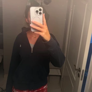 Marinblå hoodie från Polo Ralph Lauren - Marinblå hoodie från Polo Ralph Lauren med klassisk vit logga på bröstet. Tröjan har dragkedja framtill, huva och långa ärmar. Mjuk bomullskvalitet och normal passform. Perfekt för chill dagar eller när du vill ha en enkel och snygg look.