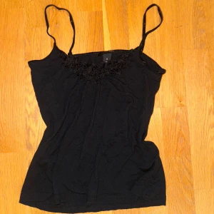 Svart linnetopp med spetsdetalj - Svart linnetopp med smala axelband och snygga spetsdetaljer längs urringningen. Toppen har en enkel, figurnära passform och är perfekt för att skapa en stilren look. Materialet känns mjukt och lätt mot huden.