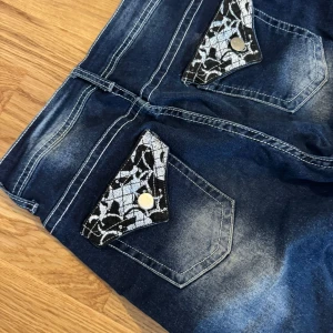 Blå bootcut jeans med spetsdetalj - Mörkblå bootcut jeans med coola ficklock bak i svart och vit spetsmönster och silvriga knappar. Jeansen har markerade sömmar och lätt slitna partier framtill för en trendig look. Perfekta för dig som vill sticka ut lite extra.