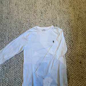 Vit långärmad tröja från Polo Ralph Lauren - Vit långärmad tröja från Polo Ralph Lauren i mjuk bomull. Klassisk rund hals och ikonisk broderad logga i marinblått på bröstet. Perfekt basic för garderoben, enkel att matcha med jeans eller shorts. Snygg och stilren design som alltid funkar.