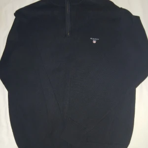 Svart stickad half zip-tröja GANT - Svart stickad tröja från GANT i storlek S med half zip och ribbade muddar. Tröjan har GANT-logga broderad på bröstet och gulddetaljer vid dragkedjan. Tillverkad i 100% bomull, perfekt för dig som gillar klassisk och stilren look.