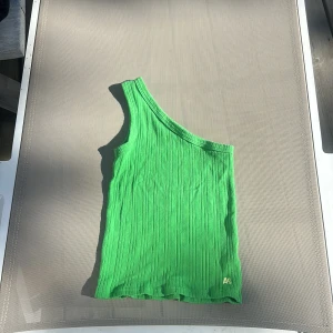 Grön oneshoulder topp AO76 - Snygg grön ribbad topp från AO76 med oneshoulder-design. Toppen har en cool asymmetrisk form och är ärmlös, med broderad logga i nederkant. Perfekt för dig som vill sticka ut med färg och stil i sommarvärmen. Ny pris ligger på 499kr