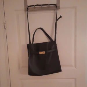 Svart axelväska med gulddetaljer - Nyskick! Stilren svart axelväska med justerbar axelrem och ett snyggt guldfärgat lås framtill. Väskan har en klassisk fyrkantig form och praktiskt handtag upptill. Perfekt för dig som gillar minimalistisk design och vill ha plats för det viktigaste. 