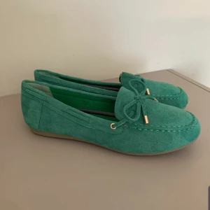 Gröna mockaloafers med rosett - Snygga loafers i grön mocka med platt sula och rund tå. Skorna har en dekorativ rosett framtill med guldfärgade detaljer och en klassisk seglarsko-look. Perfekta för dig som gillar stilrena och bekväma skor med lite extra färg.