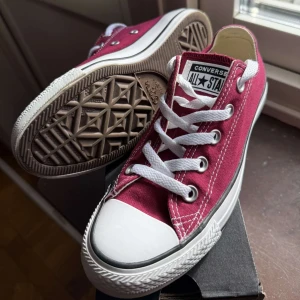 Vinröda Converse All Star sneakers - Säljer ett par klassiska Converse All Star sneakers i vinrött med vit tå och vita snören. Skorna har låg modell, rund tå och platt sula. Materialet är slitstark bomull med snygga kontrastsömmar och metallöljetter. Perfekt för dig som gillar retrostil och vill sticka ut.