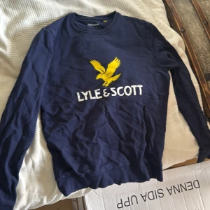 Marinblå Lyle & Scott tröja med tryck - Säljer en marinblå långärmad tröja från Lyle & Scott med stort gult fågeltryck och vit logga på bröstet. Tröjan har rund hals och är gjord i mjuk bomull, perfekt för chill dagar. Passar dig som gillar klassisk streetwear med tydlig branding.