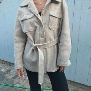 Beige kappa med bälte - Snygg beige kappa med bälte och knappar framtill. Jackan har långa ärmar och två stora fickor på bröstet. Perfekt för en stilren look.