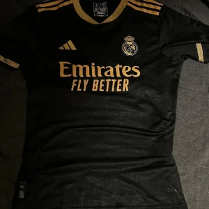 Svart Real Madrid fotbollströja - Snygg svart Real Madrid fotbollströja från Adidas med guldiga detaljer och klubbens emblem på bröstet. Tröjan har korta ärmar och är perfekt för alla fans av laget. Den har även sponsorloggan 'Emirates Fly Better' i guld på framsidan. B: 45 L: 65