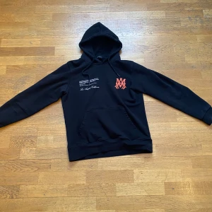 Svart hoodie från Amiri - Säljer en svart hoodie från Amiri med tryck i vitt och orange. Hoodien har en känguruficka och justerbar huva med dragsko. Perfekt för en avslappnad stil. KOSTAR 13000kr ny.
