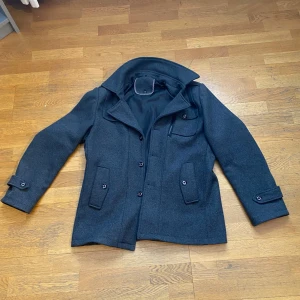 Grå kappa med knappar - Snygg grå kappa med knappar och flera fickor. Jackan har en klassisk design med krage och långa ärmar. Perfekt för kyligare dagar.