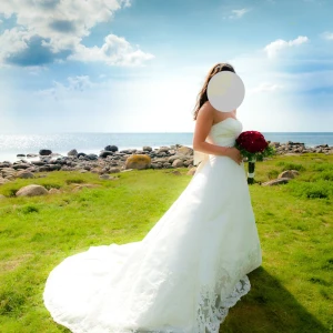 Vit axelbandslös brudklänning med spetsdetaljer - Elegant vit brudklänning med axelbandslös design och vacker spets. Klänningen har en stor rosett bak och fina broderade detaljer. Perfekt för en romantisk bröllopsdag vid havet. Rosetten går att ta av. Finns även möjlighet att ta upp släpet så att det inte drar i backen. Kemtvättad efter en användning. Underkjol ingår. Mått: Byst 94cm, midja 74cm