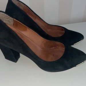 Svarta mockapumps K.Cobler  med klack - Eleganta svarta pumps i mocka med spetsig tå och klack( 9 cm). Skorna har en stilren design med en subtil gulddetalj vid tån. Perfekta för en chic look.