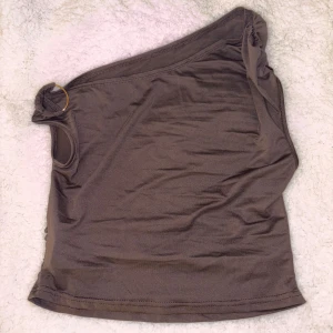 Brun oneshoulder topp - Snygg brun oneshoulder topp med en dekorativ ringdetalj vid axeln. Perfekt för en stilren och modern look. Materialet är mjukt och bekvämt.