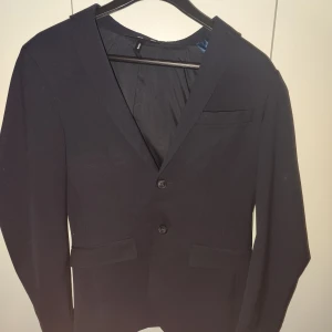 Svart kavaj med två knappar - Snygg svart kavaj från Jeansbolaget i herrstorlek S. Kavajen har en klassisk design med två knappar framtill och fickor med lock. Perfekt för både formella och semi-formella tillfällen.