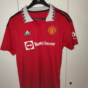 Röd Manchester United fotbollströja från Adidas - Säljer en röd Manchester United fotbollströja från Adidas med vit krage och svarta detaljer. Tröjan har korta ärmar och klubbens emblem på bröstet. Perfekt för fans som vill visa sitt stöd.
