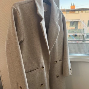 Beige kappa  - Elegant beige kappa från Stories i storlek XS. Kappan har en klassisk design med knappar framtill och långa ärmar. Perfekt för våren. Använd enstaka gånger. Bra kvalité.