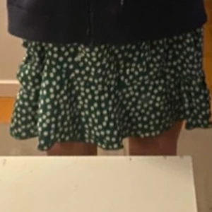 Grön blommig kjol med volang - Csupper fin grön kjol med vita blommor och volangdetaljer. Kjolen har en design med knytband i midjan som kan knytas om önskas. Perfekt för en somrig look! Bra skick. Inga fläckar och inga hål. 
