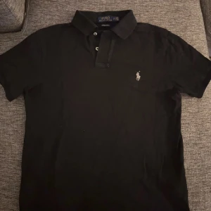 Svart pikétröja från Ralph Lauren - Säljer en klassisk svart pikétröja från Ralph Lauren med den ikoniska loggan broderad på bröstet. Tröjan har en krage och knappar framtill. Perfekt för en stilren look.