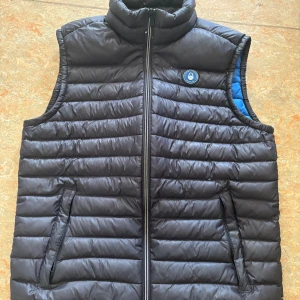 Svart dunväst med dragkedja - Snygg svart dunväst med dragkedja och hög krage. Västen har ett blått foder och två praktiska fickor framtill. Perfekt för kyliga dagar.