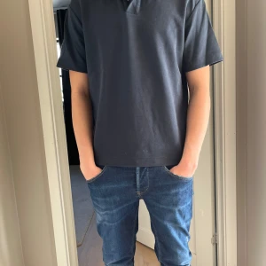 Svart kortärmad pikétröja - Säljer en stilren svart pikétröja med korta ärmar och klassisk krage. Perfekt för en avslappnad men ändå snygg look. Tröjan har knappar vid halsen för en justerbar passform.