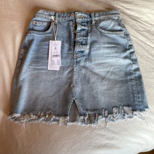 Blå jeanskjol med slitna detaljer - Snygg blå jeanskjol med slitna kanter och en liten slits framtill. Kjolen har en klassisk femficksdesign och stängs med knappar. Perfekt för en avslappnad stil. Den är helt oanvänd med prisplapp på från Gina 