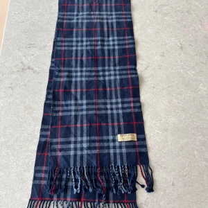 Rutig halsduk från Burberry - Snygg rutig halsduk från Burberry i blått och rött. Tillverkad i 100% lammull, perfekt för att hålla dig varm. Typ nyskick! Skriv vid frågor eller funderingar!